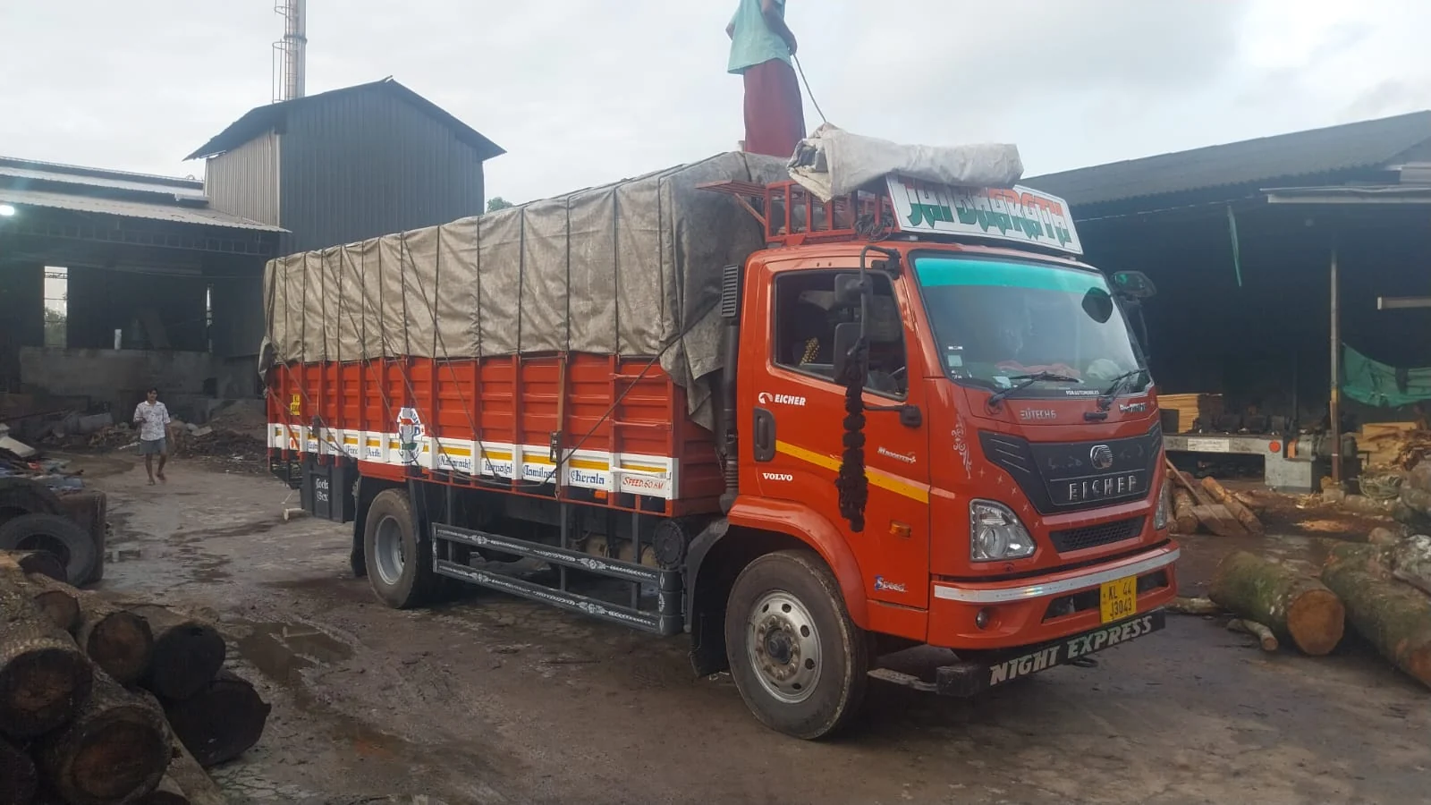 A covered red Indian lorry beside logs and industrial sheds